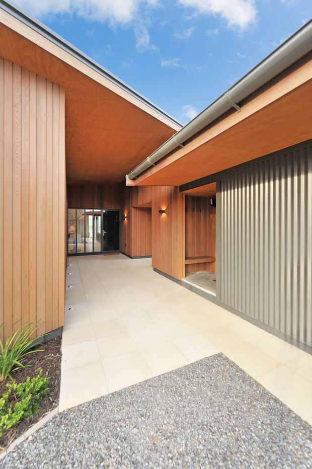 A contemporary covered entry courtyard framed by warm timber-clad walls and soffits. Pale tiled paving leads toward a glazed entrance, with a ribbed grey metal garage or service door on the right. Exposed gutters, recessed lighting, and a gravel forecourt complete the minimalist architectural composition.