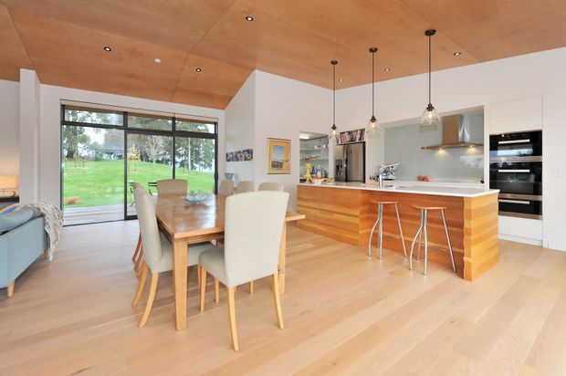 Open-plan kitchen and dining area with light timber flooring, a timber-lined ceiling, white walls, and extensive glazing overlooking a green outdoor setting. The kitchen features a large timber island with white benchtop, integrated appliances, pendant lighting, and a dining table with upholstered chairs.