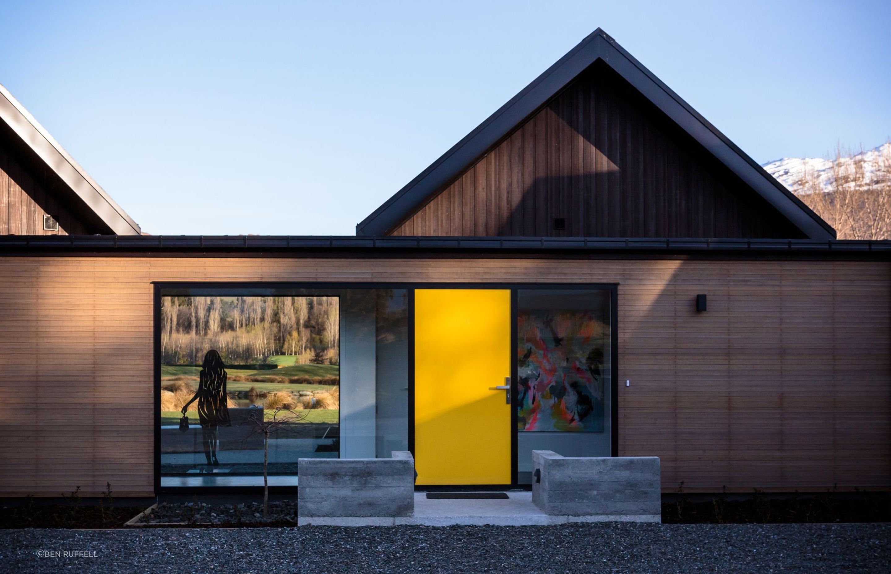 The bright yellow, aluminium front door sets up the sense of arrival and serves as a counterbalance to the mid-ground view spied through the adjacent window.