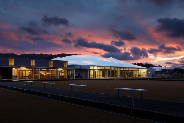 A low, modern institutional or commercial building photographed at dusk, with extensive warm-lit floor-to-ceiling glazing, dark rectangular facade volumes, and a prominent curved white roof canopy. The building fronts a large paved or sports-court-like open area with simple benches, silhouetted hills, and a dramatic pink and purple sunset sky in the background.