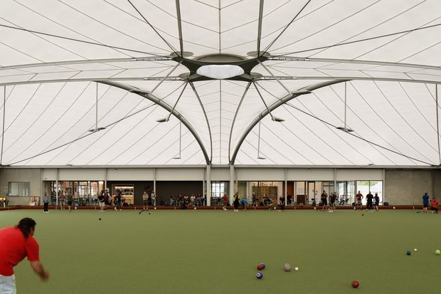 Interior view of a large indoor lawn bowls facility, with a broad green playing surface, bowls and players in the foreground, and a dramatic tensile membrane roof supported by curved structural arches and a central hub. The perimeter contains glazed rooms and spectator or club areas.
