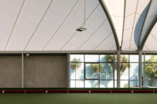 A spacious indoor sports or recreation facility with a translucent tensile canopy roof supported by curved steel columns and beams. The far wall combines raw concrete panels with large black-framed glazed sections, opening views to palm trees outside. A red horizontal rail or barrier runs along the lower wall, and a green playing surface occupies the foreground. The space is brightly lit with a clean, utilitarian architectural character.