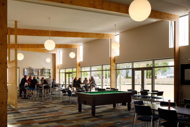 A bright, expansive communal interior with exposed timber beams and columns, large floor-to-ceiling windows, suspended spherical pendant lights, café-style tables and chairs, and a central billiards table. A group dining or social area is visible toward the rear, with a patterned multicoloured carpet throughout.