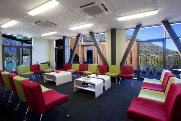 A bright contemporary commercial lounge or reception area furnished with clusters of colourful red and lime-green armchairs around white coffee tables. Floor-to-ceiling glazing provides expansive mountain views, while exposed diagonal structural bracing, timber-clad columns, dark carpet, and a suspended grid ceiling with recessed air-conditioning and fluorescent lighting define the space.