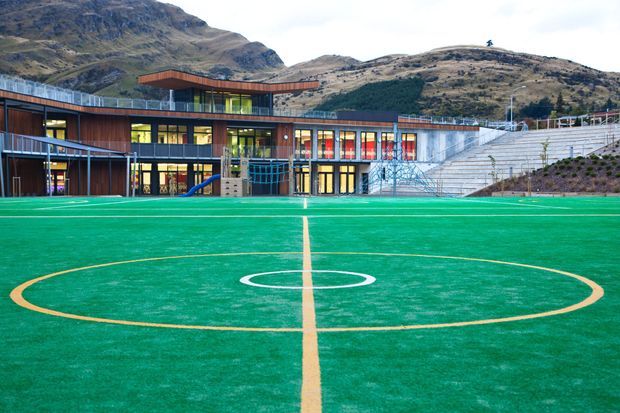 Exterior view of a contemporary multi-storey school or community sports facility set against steep mountainous terrain. The building features extensive glazing, dark and warm timber-toned cladding, projecting roof forms, outdoor play equipment, broad concrete spectator steps, and a bright green marked sports field in the foreground.
