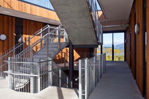 A multi-level contemporary building circulation space with exposed concrete walkways and staircases, vertical timber wall cladding, and silver metal balustrades. The open corridor frames a distant mountainous landscape through full-height glazing.