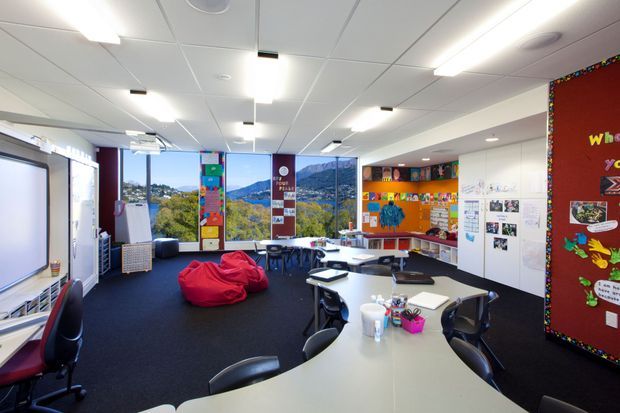 A bright contemporary classroom or learning space with large windows overlooking a mountainous landscape. The room contains clustered white tables, black chairs, a red beanbag, interactive teaching boards, colourful student displays, pinboards, shelving, and extensive white cabinetry. Suspended fluorescent lighting and acoustic ceiling tiles create a practical educational interior.
