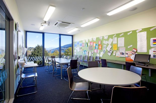 A bright, contemporary shared classroom or meeting space with round and rectangular tables, stackable chairs, desks with computers, and a large green pinboard wall covered in notes and displays. Floor-to-ceiling glazing provides views of a leafy mountainous landscape. Linear fluorescent-style ceiling lights and a carpeted floor complete the practical institutional interior.