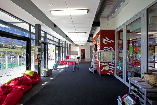 A bright contemporary educational or community interior corridor with full-height glazing on the left, dark carpet flooring, a suspended acoustic ceiling with recessed and rectangular fluorescent lighting, and exposed dark structural beams. The space includes colourful informal seating, display boards, shelves, and glass-fronted rooms or offices along the right. A landscaped sports or recreation area is visible through the windows.