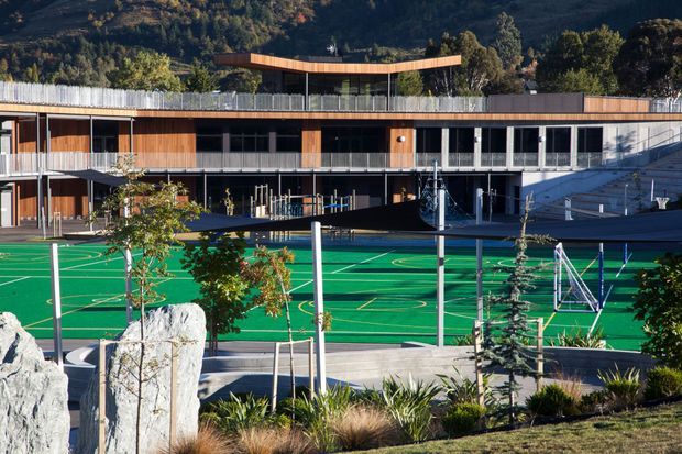 Exterior view of a contemporary multi-level school or institutional building overlooking bright green outdoor sports courts, with timber-clad façades, extensive glazing, covered walkways, planted landscaping, and a wooded hillside backdrop.