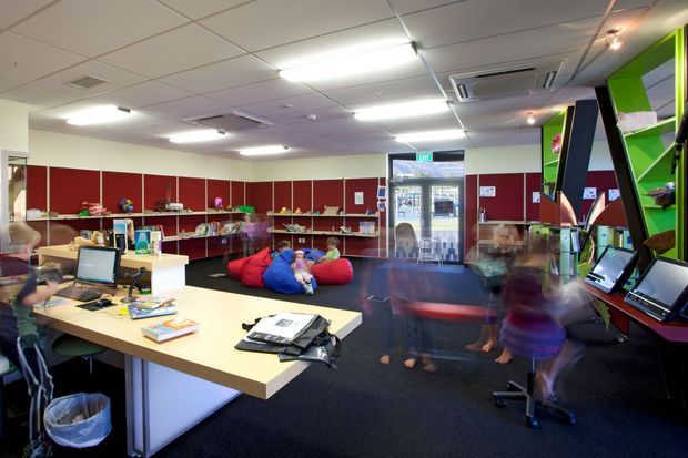 A brightly lit commercial interior resembling a school library, learning commons, or youth activity space. The room contains a large reception or work desk with computers, extensive low display shelving, colourful beanbag seating, and a dark carpeted floor. Red and lime-green display areas provide bold accents, while a glazed entrance is visible at the rear. People are present but are not the focus.