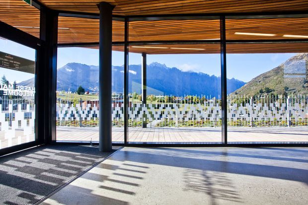 A spacious, glazed architectural interior with full-height black-framed windows overlooking a dramatic mountain landscape and vineyard-like grounds. The room features a polished concrete floor, exposed dark structural columns, and a slatted timber ceiling with integrated linear lighting. Decorative geometric frosting covers sections of the glazing, while strong sunlight creates patterned shadows across the floor.