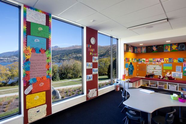 Bright, colourful primary school classroom with large windows overlooking a mountainous landscape and water. The room features student artwork and displays, colourful wall graphics, built-in storage and shelving, a collaborative kidney-shaped table with chairs, and dark carpet flooring.