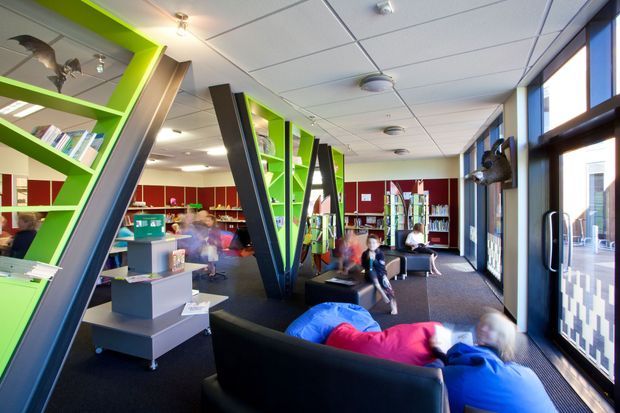 Bright contemporary library or reading lounge with vivid lime-green built-in shelving, angled dark structural frames, red accent walls, books, display units, and large windows along one side. The space includes soft seating and children or young people using the area, with a suspended acoustic ceiling and dark carpet flooring.