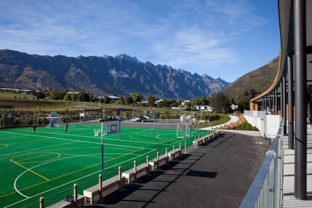 A wide exterior view of a school or community sports facility beside a large green multi-court basketball and netball court. The paved spectator walkway and building edge run along the right, while a dramatic rugged mountain range and rural valley landscape form the background.