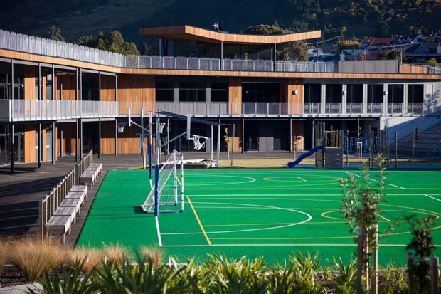Exterior view of a contemporary multi-level educational or recreational building surrounding a bright green sports court. The structure features extensive glazing, warm timber cladding, dark metal framing, balconies and railings, with a curved roof canopy and play equipment visible at ground level. Landscaping occupies the foreground, with wooded hills in the background.