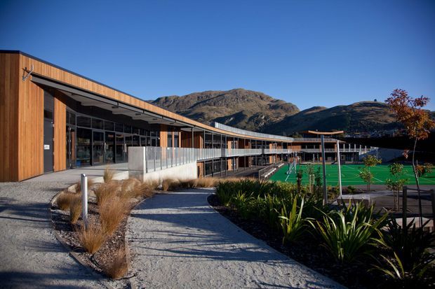 Exterior view of a contemporary, low-rise building with warm vertical timber cladding, extensive glazing, and a long covered walkway overlooking landscaped grounds and green sports courts. Rugged mountains rise in the background under a clear blue sky.
