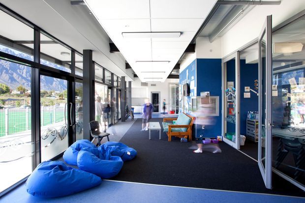 Bright contemporary institutional interior corridor or shared lounge with full-height glazing, dark carpet flooring, white tiled ceiling, exposed services, blue accent walls, and glass-panelled rooms. Blue beanbags and timber-framed lounge chairs provide informal seating. A fenced sports court and mountainous landscape are visible through the glazing, while people are blurred in motion.