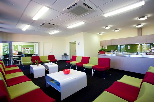 A bright, contemporary commercial lounge or reception area with clusters of lime-green and red upholstered seating arranged around white modular coffee tables. The room has dark carpet, pale walls, a suspended acoustic ceiling with extensive linear fluorescent lighting and recessed downlights, and a long built-in reception or kitchenette counter with overhead cabinetry and a reflective backsplash. A small meeting or work area is visible through the glazed partition at the rear.
