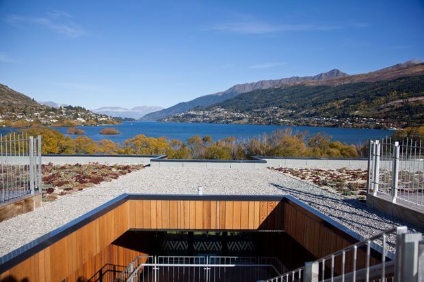 A rooftop terrace or upper-level outdoor area overlooks a blue alpine lake surrounded by forested mountains and hillside homes. The foreground features a broad gravel-covered roof terrace with low planted beds, metal safety railings, and a recessed stairwell lined with warm timber cladding.