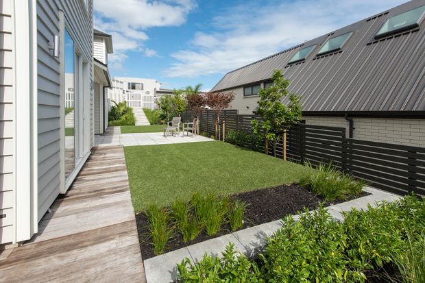 A landscaped residential courtyard or side garden between contemporary buildings. The space includes a timber boardwalk along a grey weatherboard-clad building, a compact lawn, dark garden beds with ornamental grasses and shrubs, concrete stepping slabs, and a black horizontal-slat fence. A neighbouring building has a steep grey metal roof with skylights and white brick walls. The scene is bright and sunny with a clean, contemporary outdoor design.