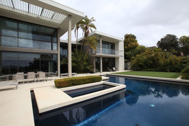 Modern multi-level residential home with extensive floor-to-ceiling glazing, white horizontal cladding, broad flat roof overhangs, and glass balcony balustrades. A large swimming pool and raised rectangular spa dominate the foreground, with a manicured lawn and tropical landscaping surrounding the house.