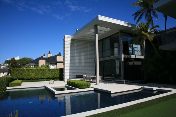Exterior view of a contemporary two-storey residence with a flat roof, expansive dark-framed glazing, a light horizontal-clad feature wall, covered outdoor entertaining terrace, manicured hedges, and a geometric swimming pool with raised square water feature. Palm trees and neighbouring homes are visible in the background.