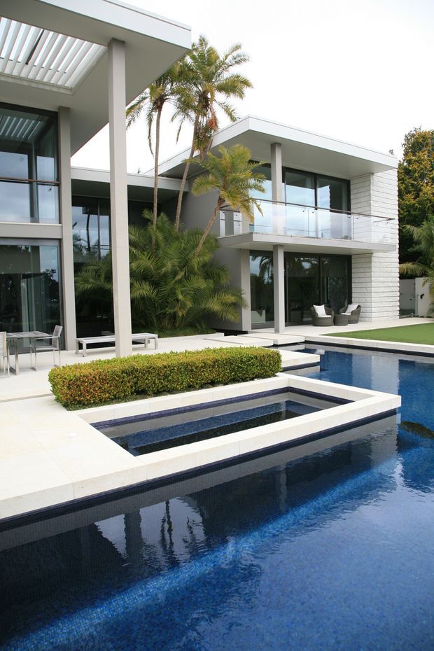 Exterior view of a large contemporary two-storey residence arranged around a landscaped pool courtyard. The home features crisp white rendered and masonry walls, flat roof planes, expansive dark-framed glazing, glass balcony balustrades, slender structural columns, and a covered outdoor entertaining area. A deep blue swimming pool with stepped raised sections dominates the foreground, surrounded by pale paving, clipped hedging, palms, and low outdoor furniture.