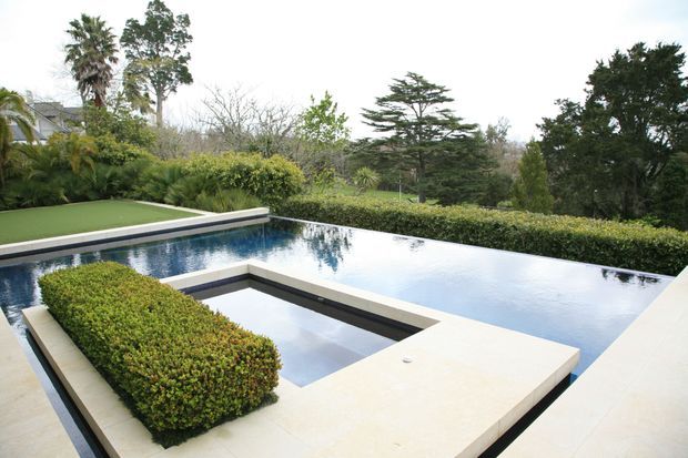 A modern geometric outdoor pool area with a reflective infinity-style pool, pale stone coping and broad paved surrounds. A smaller recessed square section is integrated into the pool design, with neatly clipped hedges and manicured greenery framing the water. Mature trees and landscaped gardens form the background.