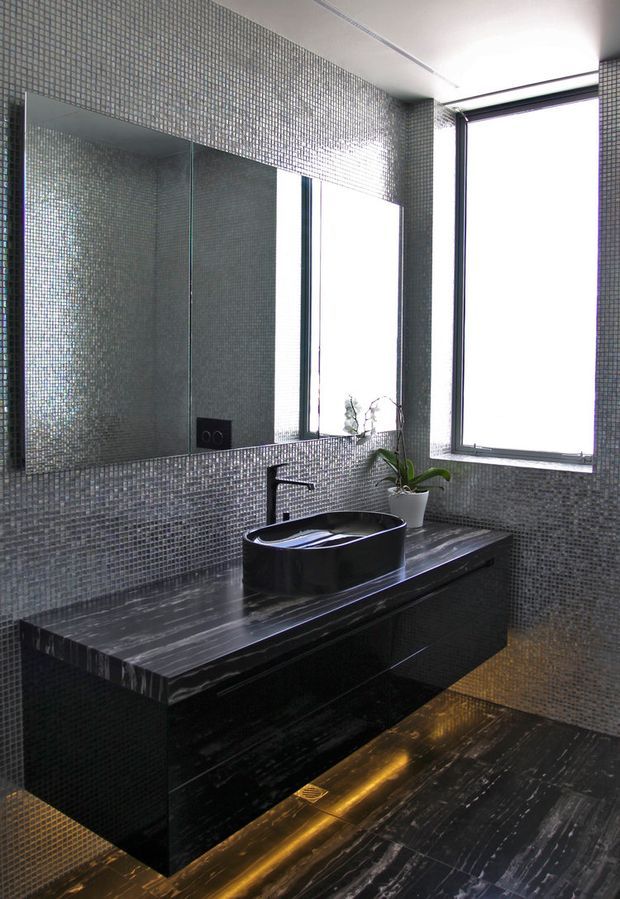 A dramatic contemporary bathroom featuring dark grey mosaic tile walls and floor, a large frameless mirror, black stone vanity with integrated cabinetry, a black vessel basin and minimalist black tapware. Tall windows provide strong natural light, while concealed warm lighting accents the floor and vanity.