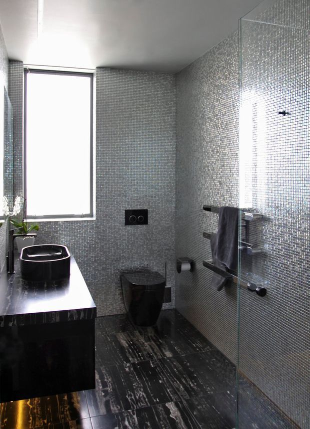 Compact contemporary bathroom with dark stone-look flooring, black floating vanity and vessel basin, wall-hung black toilet, extensive small grey mosaic wall tiling, a frameless glass shower screen, and chrome towel rails and fittings. Natural light enters through a tall window.