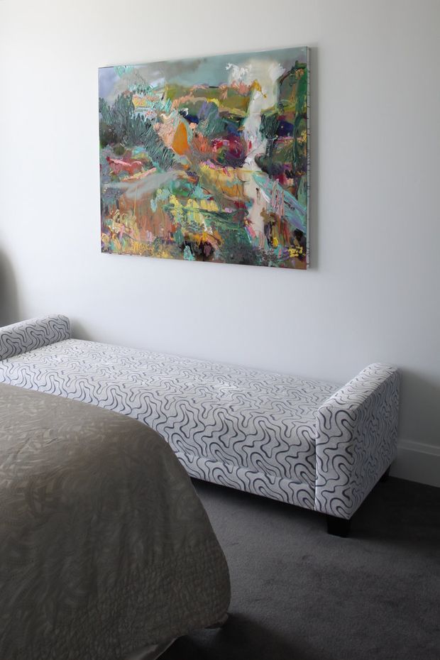 A quiet bedroom corner with a low upholstered chaise or daybed positioned along a pale wall. The daybed is covered in a white fabric with an organic black squiggle pattern and has a rounded bolster-style end. A large colourful abstract painting hangs above it. The foreground shows part of a bed with a muted grey quilt, while dark grey carpeting and simple white skirting complete the room.