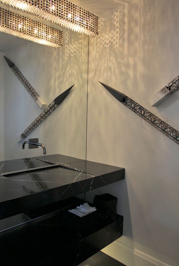 A dramatic, luxurious bathroom vanity viewed through a large mirror. The space features a floating black stone vanity with integrated rectangular basins, a chrome wall-mounted faucet, pale walls, and sculptural geometric wall accents. Crystal-like rectangular pendant lights cast patterned reflections across the walls, creating a high-contrast, glamorous atmosphere.