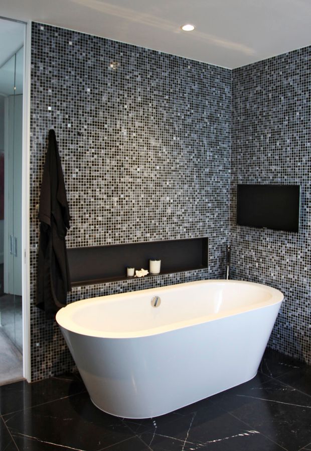 Contemporary bathroom featuring a freestanding white bathtub, dark floor tiles, and walls clad in small grey, black, and silver mosaic tiles. A wall-mounted black television, recessed shelf, shower area, and dark towel add to the sleek monochromatic design.