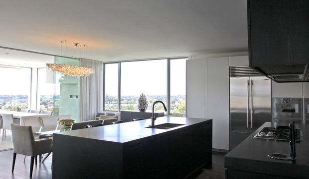 A refined contemporary open-plan kitchen with a large dark island, integrated sink and black tap, dark cabinetry, stainless-steel appliances, and expansive floor-to-ceiling glazing. A dining area and sculptural pendant light are visible beyond the kitchen, with an elevated city and landscape outlook.