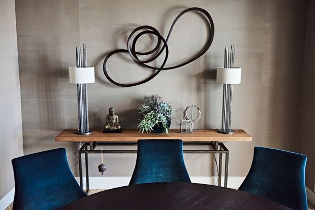 A refined dining area with a round dark dining table and three deep blue upholstered chairs. Behind the table is a slim console with a warm timber top and dark metal frame, flanked by tall sculptural lamps. A large abstract looping wall sculpture and small decorative objects create a sophisticated feature wall. The room has neutral taupe walls and a calm, contemporary mood.