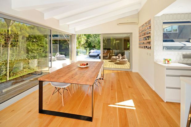 Bright open-plan dining area with a long timber dining table and modern chairs, honey-toned timber flooring, extensive glazing, and exposed white ceiling beams. A compact white kitchen is visible to the right, while the glazing opens toward a leafy outdoor setting and reflects an adjacent living area.