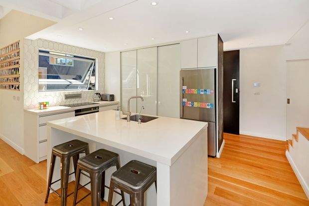 Bright contemporary kitchen with a large white island and integrated sink, three metal bar stools, white cabinetry, stainless-steel refrigerator, patterned hexagonal tile splashback, timber flooring, and a built-in open bookshelf. A dark timber-framed doorway and stair edge are visible in the background.
