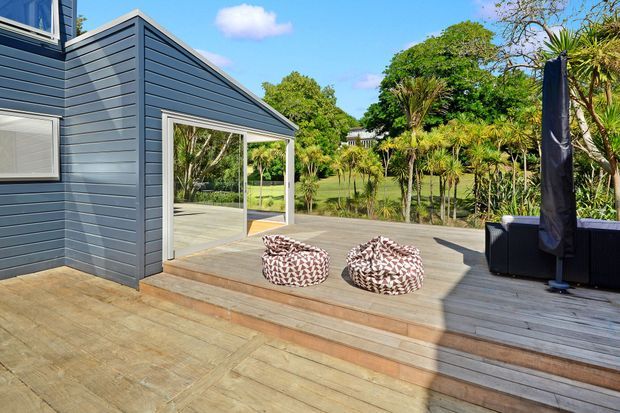 Sunny exterior view of a contemporary dark-grey weatherboard-clad building opening onto a broad timber deck. The deck has two patterned low seating poufs and an outdoor lounge area, with lush landscaped planting beyond. Large sliding glass doors connect the interior to the deck.