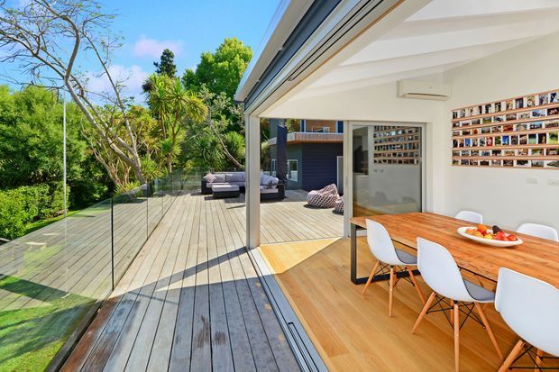 Bright contemporary dining and outdoor entertaining area opening through large sliding glass doors onto a spacious timber deck. The interior has white walls and ceiling beams, timber flooring, a built-in wall display, and a timber dining table with white modern chairs. The deck features frameless glass balustrading and overlooks lush landscaped greenery.