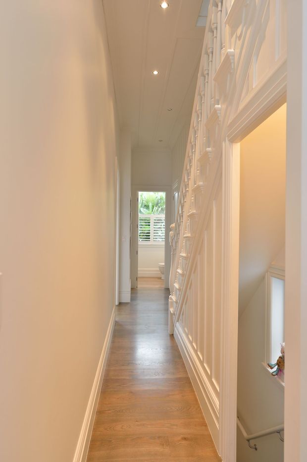 A narrow, elongated interior hallway with warm white painted walls and ceiling, recessed downlights, and a medium-toned timber floor. The right side features elaborate white traditional-style wall panelling and trim around openings, including a view toward a stairwell. A bright window at the far end looks onto greenery and a horizontal railing. The space is softly lit, refined, and distinctly traditional in detailing.