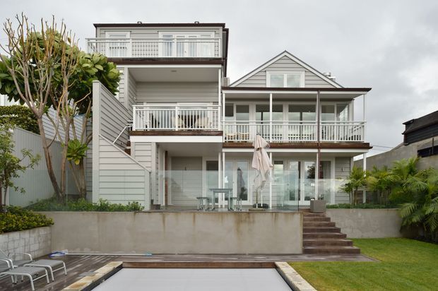 Rear exterior view of a multi-storey residential house with a gabled main roof, upper-level balconies, white railings, grey horizontal cladding, extensive glazing, and a raised landscaped terrace. A partially visible swimming pool occupies the foreground, with stairs connecting the lawn and terrace.