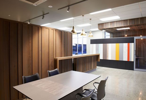 A contemporary commercial meeting or collaboration space with a large rectangular table and office chairs in the foreground. The room features extensive dark timber wall panelling and a matching partial-height divider, polished concrete or terrazzo-like flooring, suspended pendant lights, track lighting, and a suspended acoustic ceiling. A bold black-and-white partition with vertical yellow, orange, and red panels adds a graphic accent, while glazed office areas are visible beyond.