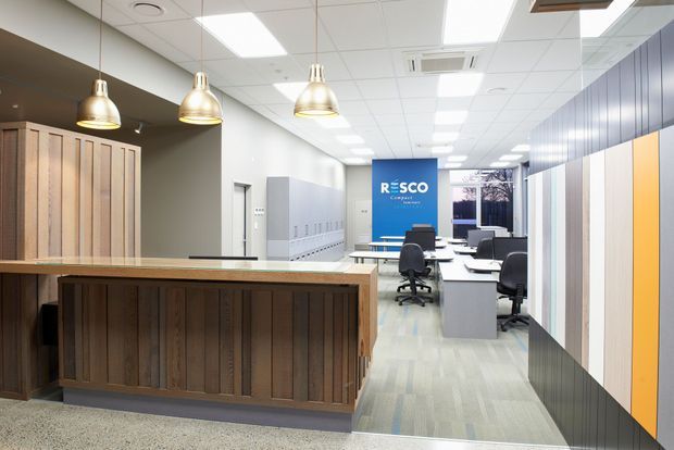 A bright contemporary commercial office reception and open-plan workspace. The foreground features a timber-clad reception counter with a pale stone or solid-surface benchtop and three metallic pendant lights. Behind it are rows of desks and office chairs, lockers, acoustic or decorative wall panels, a blue branded partition, and a suspended grid ceiling with recessed fluorescent lighting and air-conditioning.