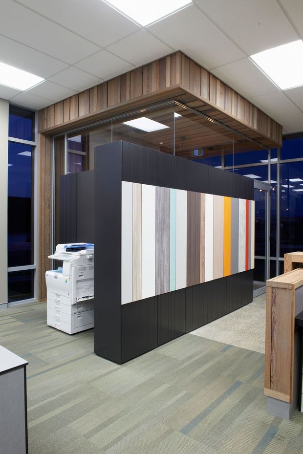 A modern commercial office interior with a freestanding black display and partition wall featuring vertical samples in timber, white, grey, pale blue, yellow and red. The space has glass partitions, timber-lined framing and ceiling soffits, blue-tinted glazing, striped carpet, fluorescent-style recessed ceiling panels, a printer, and built-in timber joinery.