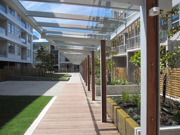 A bright landscaped pedestrian courtyard within a multi-storey contemporary development. A long timber-look walkway runs beneath a glazed, steel-framed canopy supported by slender timber-clad posts. Raised planted garden beds, lawn, timber screening and modern apartment or commercial façades line both sides, creating a sheltered outdoor circulation space.