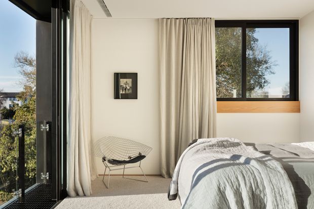 Bright, contemporary bedroom with a low-profile bed dressed in white textured bedding, full-height cream curtains, pale carpet, and a black-framed window with a timber sill. A sculptural wire lounge chair and small framed artwork add restrained detail. Glazing at the left opens to an exterior balcony or outdoor view.