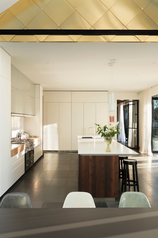 Bright, spacious contemporary kitchen viewed from an adjacent dining area. The room features white flat-panel cabinetry, a large white island with integrated sink and seating, dark timber island panelling, dark tiled flooring, stainless-steel appliances, and full-height glazing with sheer curtains. A distinctive gold-toned geometric ceiling feature with dark linear detailing spans the upper portion of the image. Natural light creates a refined, clean atmosphere.