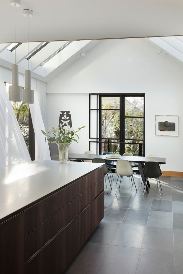 Bright, open-plan kitchen and dining area with a large dark timber island topped in a pale solid surface, a long dining table, and a mix of white and dark modern chairs. The room has a high pitched ceiling with multiple skylights, expansive black-framed glazing, sheer white curtains, grey tiled flooring, pendant lights, and restrained contemporary artwork. Natural daylight creates an airy, refined atmosphere.