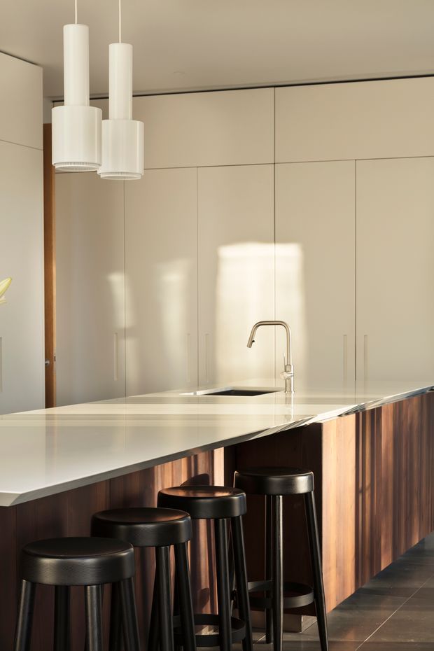 A refined contemporary kitchen centered on a long island with integrated sink and brushed metal tap. The island has a pale stone benchtop and dark vertically grained timber-clad base, paired with four black leather stools. Full-height warm off-white cabinetry provides extensive concealed storage, while simple white cylindrical pendant lights hang above. Strong sunlight creates bright reflections across the cabinetry and benchtop.